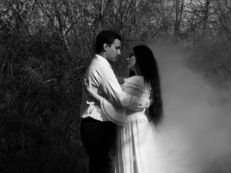 A young man and a young woman in a white dress embrace next to a mistly lake