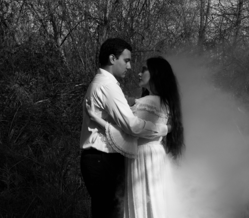 A young man and a young woman in a white dress embrace next to a mistly lake