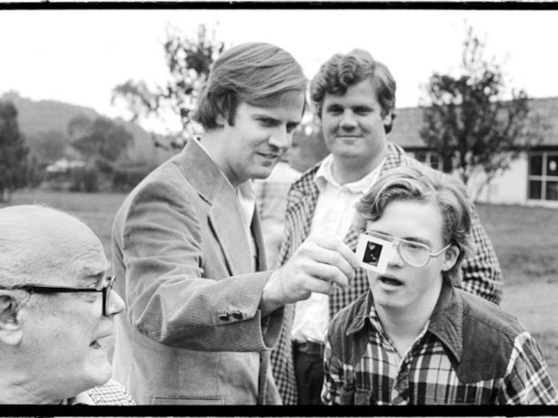 Joe stands showing his brother John a negative of one of his photos while his other brother and father look on.
