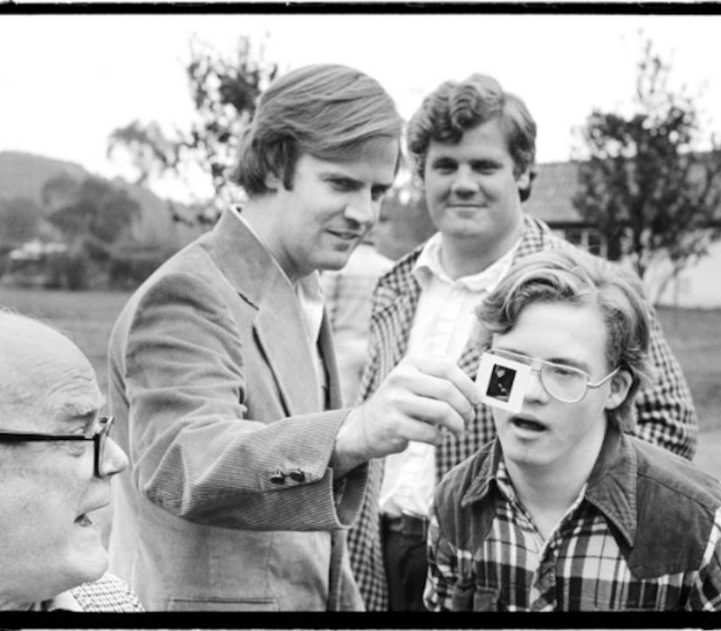 Joe stands showing his brother John a negative of one of his photos while his other brother and father look on.